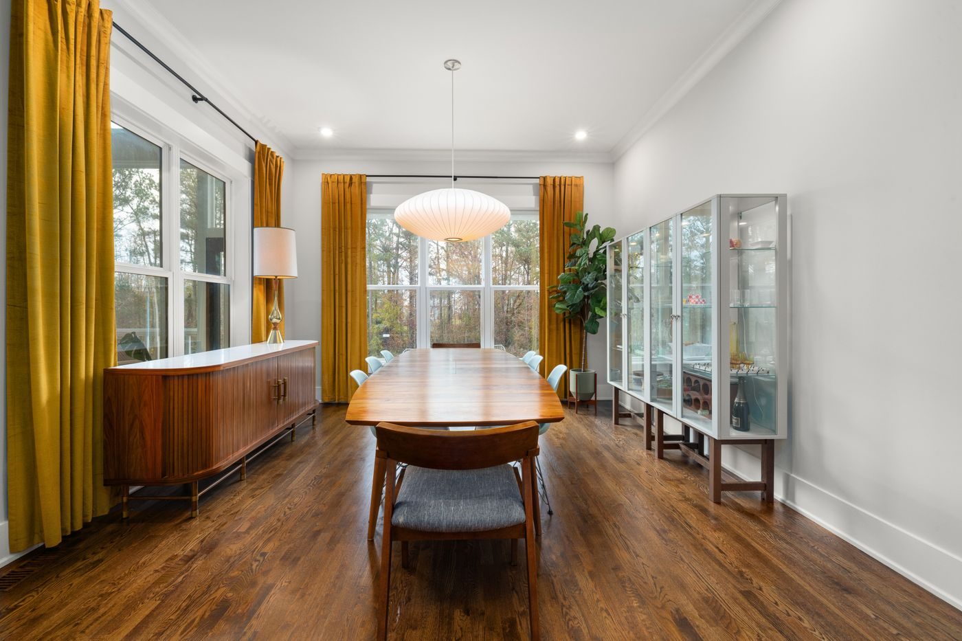 A dining room with a wood credenza, long table and warm curtains
