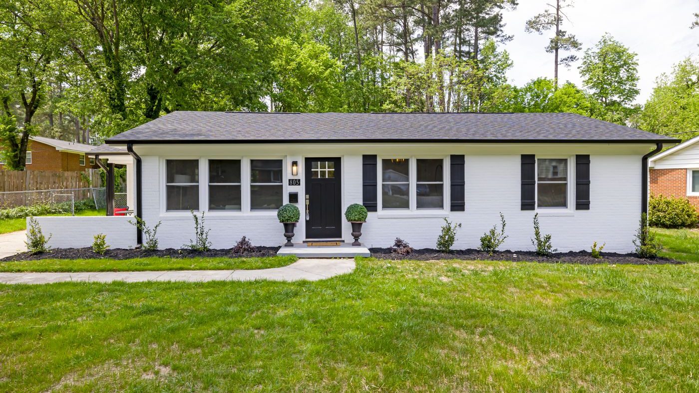 A white-painted brick ranch house with a deep lawn and tall pines behind it